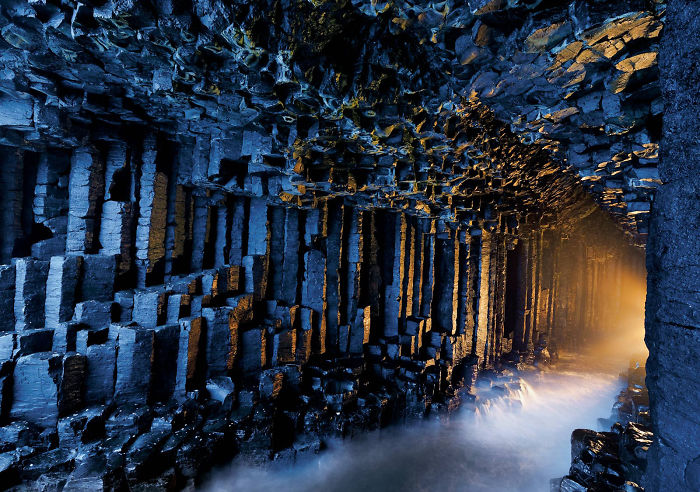 Hexagonal basalt columns inside a beautiful cave illuminated by sunlight with misty water flowing through the rocky interior.