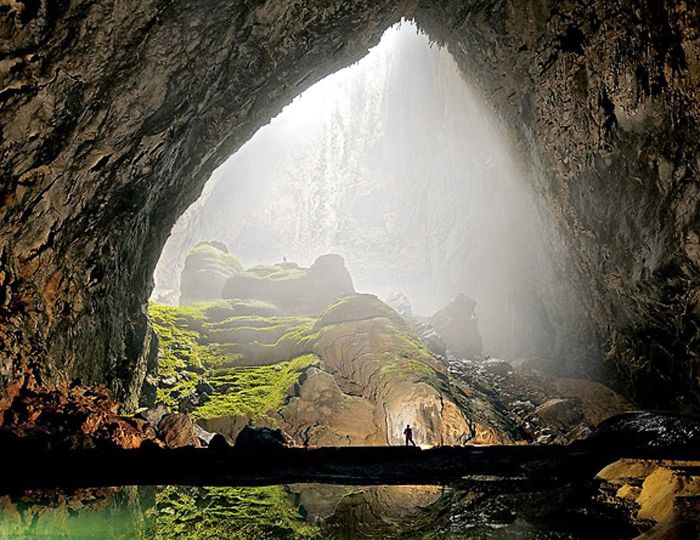 Interior view of a beautiful cave with moss-covered rocks and a person standing near its illuminated entrance.