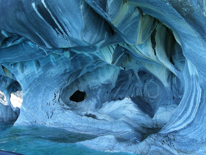 Intricate patterns and smooth formations inside a beautiful cave illuminated by blue light above clear water.