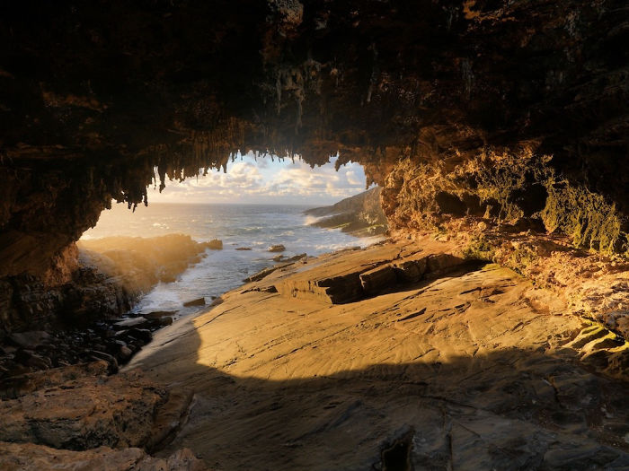 View of a beautiful cave opening with sunlight illuminating rocky interior and ocean waves outside at sunset.