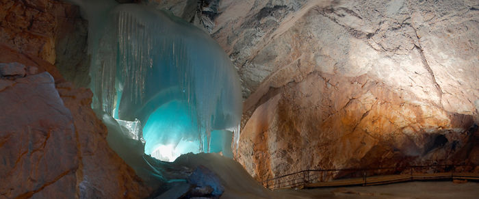 Panoramic view of a beautiful cave interior featuring large blue ice formations and rocky walls with a walkway.