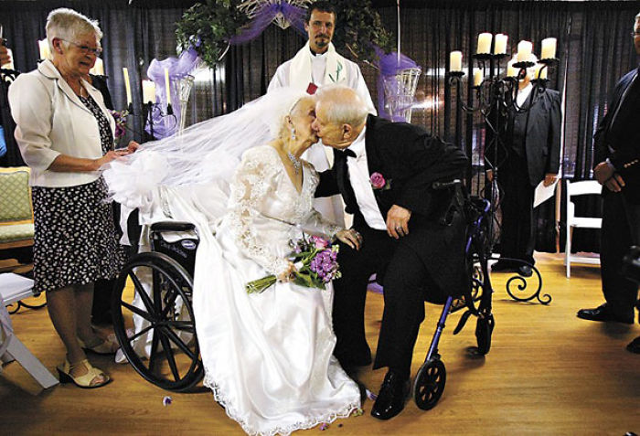 Elderly couple in wedding attire kissing, both in wheelchairs, celebrating love and fun at any age.