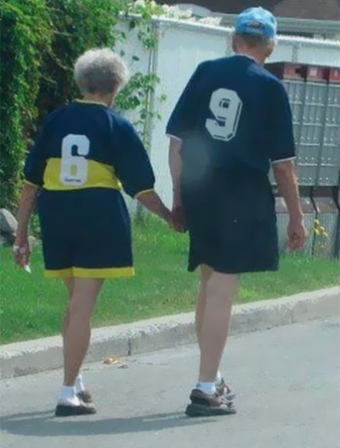 Elderly couple walking hand in hand, wearing matching jerseys with numbers on the back, enjoying a leisurely day outdoors.