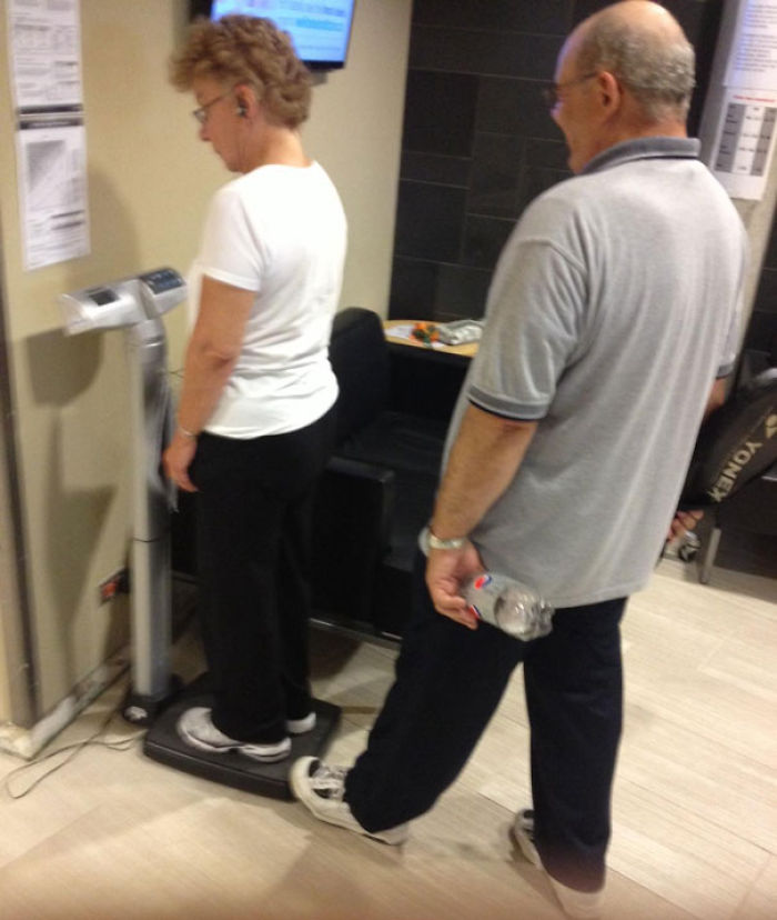Elderly couple having fun at the gym while using a scale together; man playfully holds a bottle.