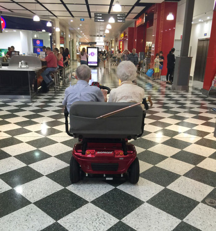 Elderly couple enjoying a ride in a shopping mall on a red mobility scooter.