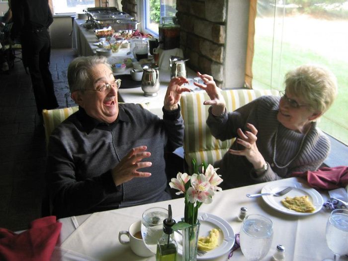Elderly couple having fun at a dining table, laughing and playfully gesturing to each other.