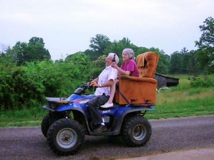 Elderly couple enjoying a fun ride on an ATV with a sofa chair attached, surrounded by greenery.