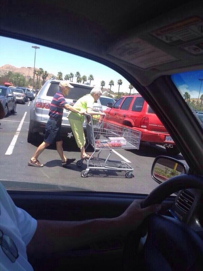 Elderly couple having fun in a parking lot, playfully riding a shopping cart under the sun.