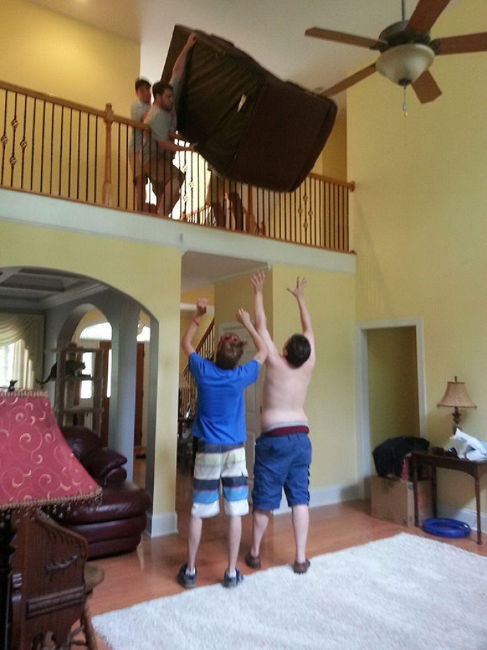 Men balancing a large couch on a balcony edge while others below prepare to catch, demonstrating risky behavior.