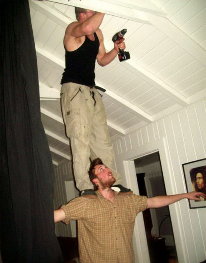 Two men performing a precarious balancing act, with one standing on the other's shoulders while using a drill.
