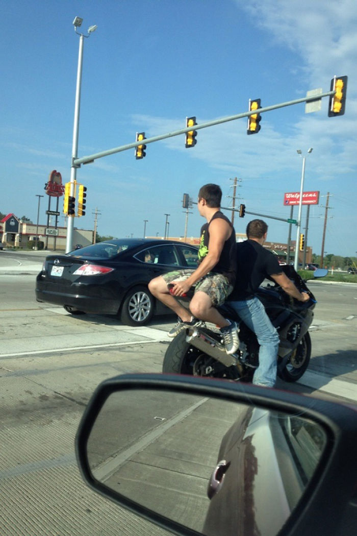 Two men on a motorcycle at a traffic light, one sitting on handlebars; illustrates reasons women may live longer than men.