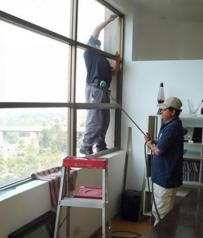 Two men cleaning high windows using a ladder inside an office, demonstrating risky behavior linked to men's shorter lifespan.