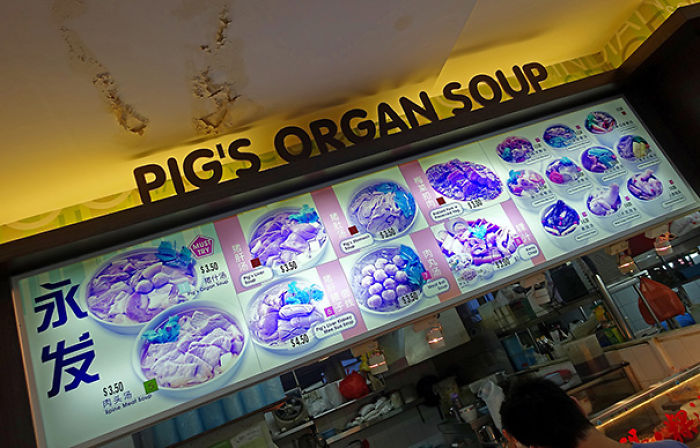 Funny menu translation fail showing "Pig's Organ Soup" with various dishes in a brightly lit food stall.