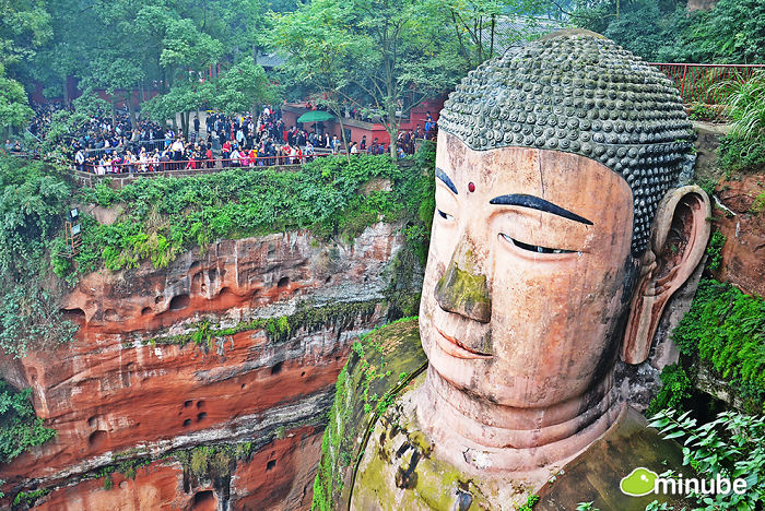Large stone Buddha statue carved into cliff surrounded by greenery and tourists, popular travel attraction in China.