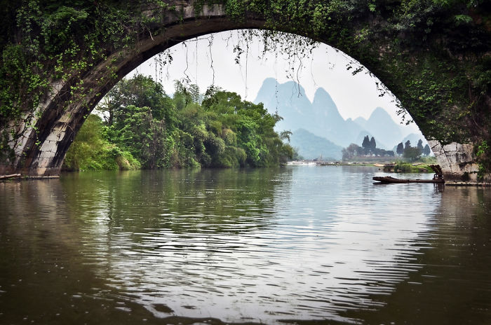 Ancient stone bridge arching over a river with lush greenery and misty mountains in the background showcasing China travel attractions.