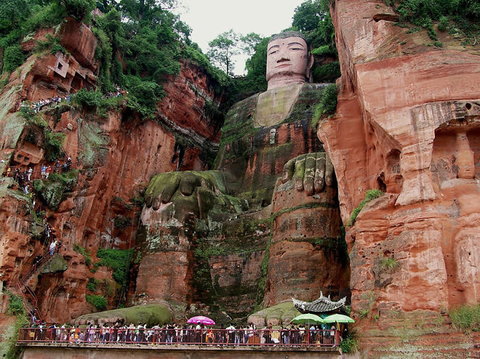 Large ancient Buddha statue carved into red cliffs surrounded by tourists, showcasing reasons to visit China travel landmarks.