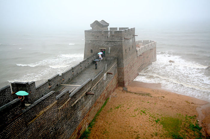 Ancient Great Wall section along the coast with visitors holding umbrellas on a foggy day in China.