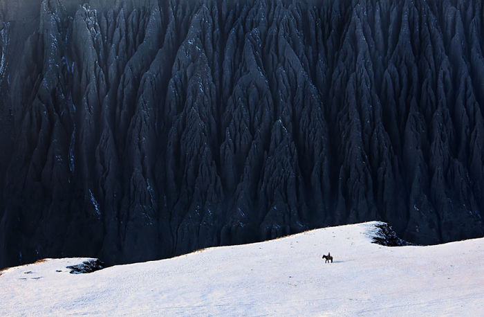 A lone rider on horseback crosses snowy terrain with dramatic dark mountains in the background in scenic China landscape.