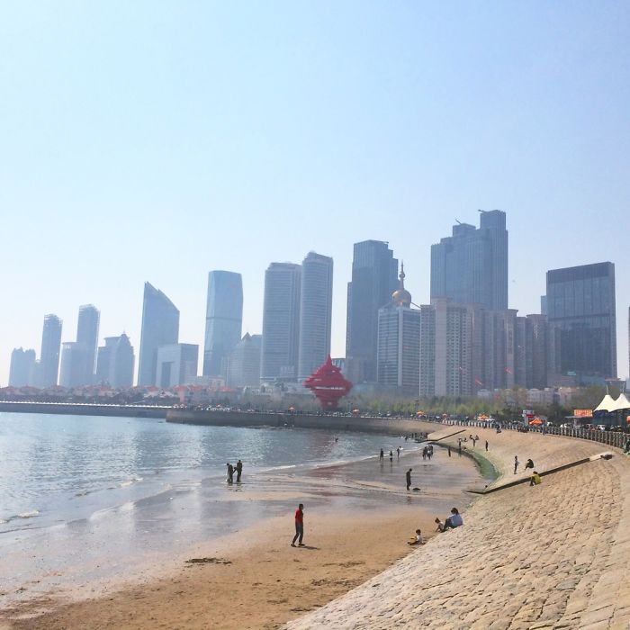 Coastal view of a modern city skyline in China with people walking along the beach and waterfront promenade.