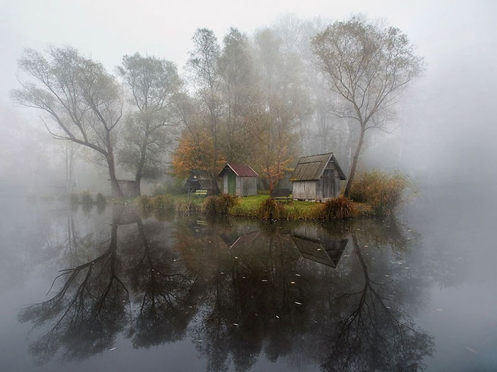 Mist-covered lake with small wooden huts and autumn trees reflecting in the water, featured in National Geographic photos.