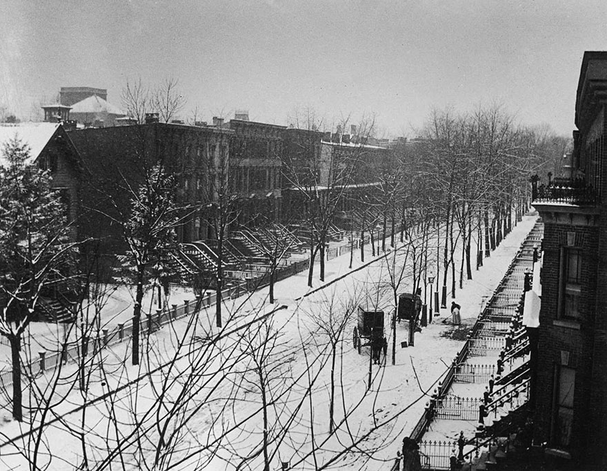 A Women On A Brownstone-Lined Street Sweeps The Sidewalk Near Where Two Horse-Drawn Buggies Are Parked In Brooklyn