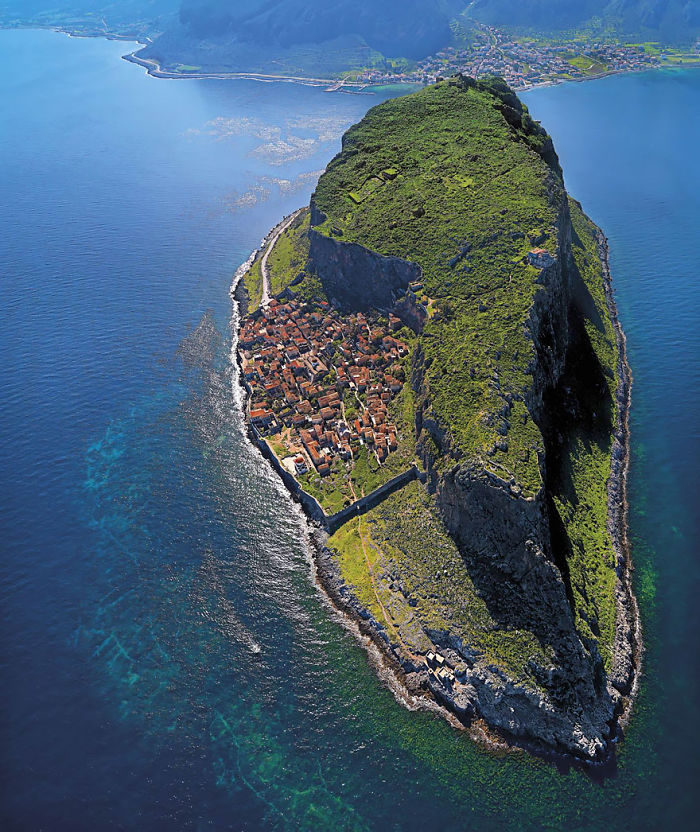 Aerial view of a fairy tale village on a rocky island with lush greenery and a surrounding sea.