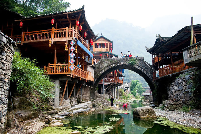 Charming village scene with wooden houses, a stone bridge over a stream, and people enjoying the fairy tale village ambiance.