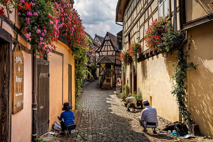 Fairy tale village street with colorful flowers and artists painting, showcasing charming architecture.