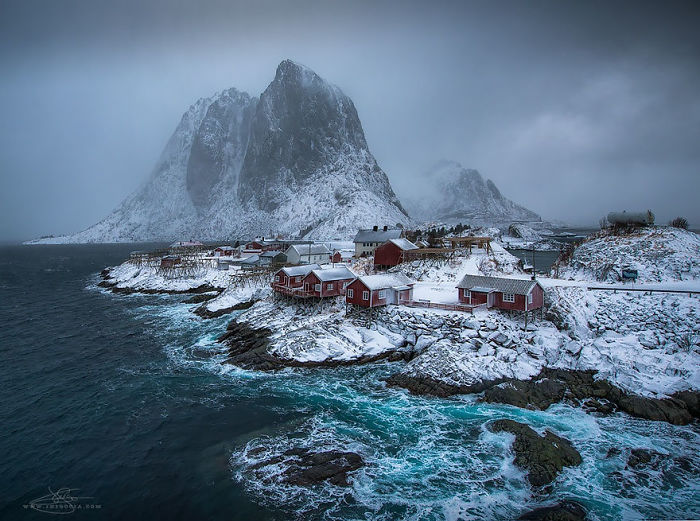 Snow-covered fairy tale village by the sea, with rugged mountain backdrop and red cabins creating a picturesque scene.