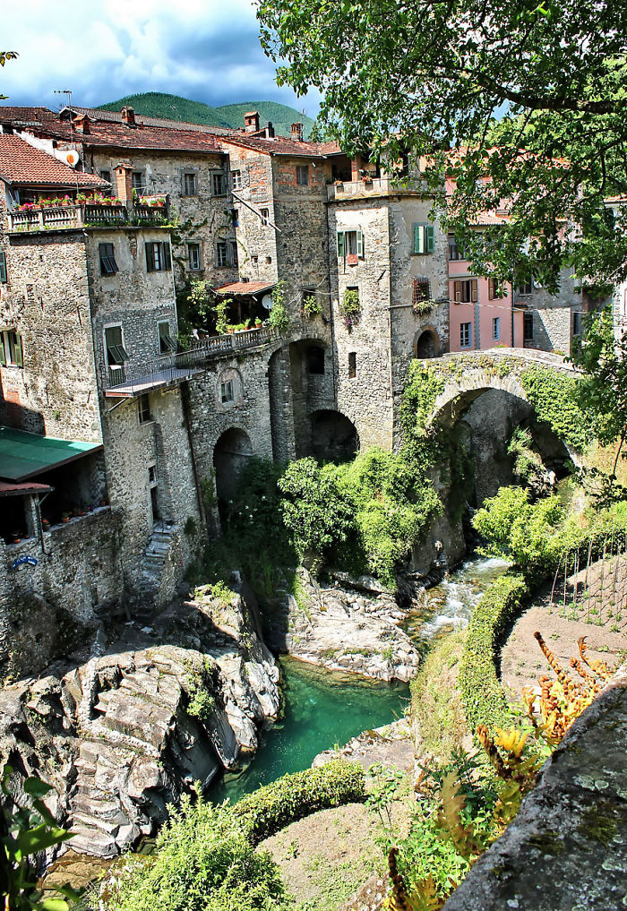 Fairy tale village with stone buildings and a lush, green river gorge under a stone bridge.