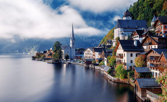 Fairy tale village by a serene lake with misty mountains in the background, showcasing charming architecture.