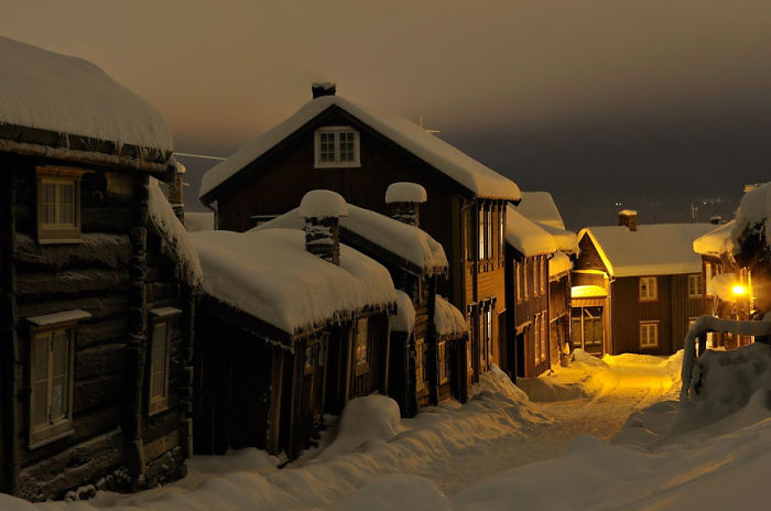 Snow-covered fairy tale village at dusk, with wooden houses and glowing streetlights.