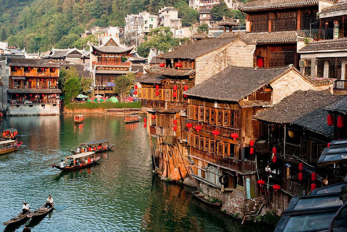 Fairy tale village with traditional wooden buildings and boats on a river, surrounded by lush greenery.