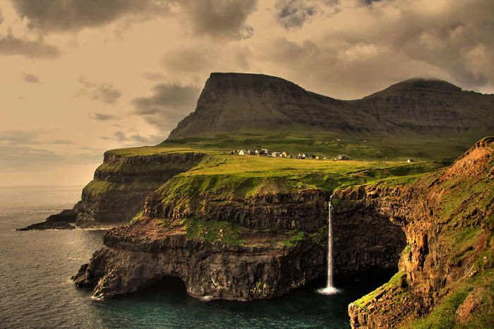 Fairy tale village perched on cliffs with a waterfall cascading into the ocean below, set against rugged mountains.
