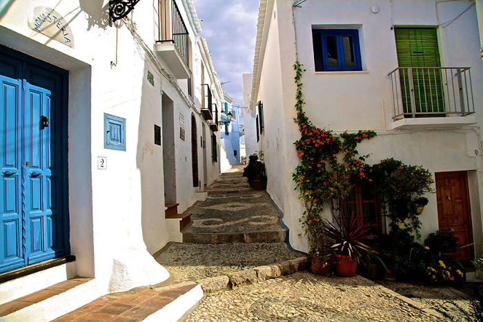 Charming fairy tale village street with white buildings, blue doors, and vibrant plants.