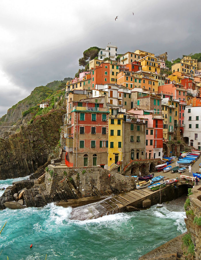 Colorful fairy tale village buildings by the sea, nestled on a rocky cliff under a cloudy sky.