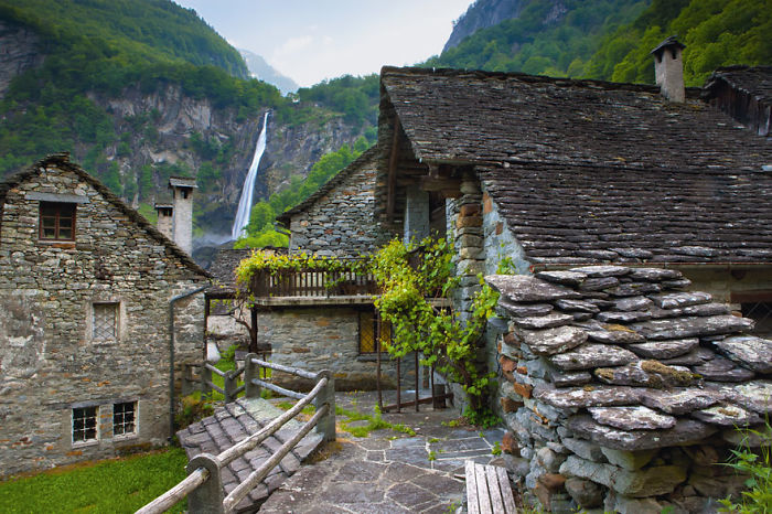 Fairy tale village with stone houses and a waterfall in the background, surrounded by lush greenery.