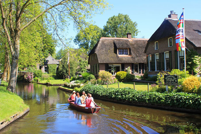 A charming fairy tale village with people boating along a canal, surrounded by lush greenery and traditional houses.