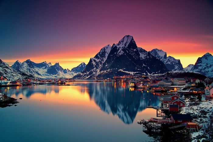 Fairy tale village by a serene lake with mountains and a vibrant sunset sky in the background.