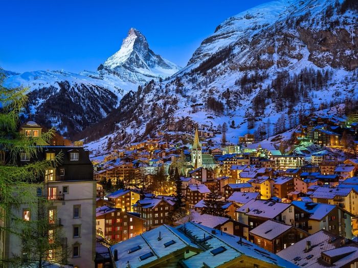 Fairy tale village with snow-covered rooftops and a mountain backdrop at twilight.