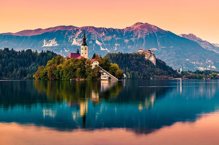 Fairy tale village by a serene lake with a church and mountains in the background at sunset.