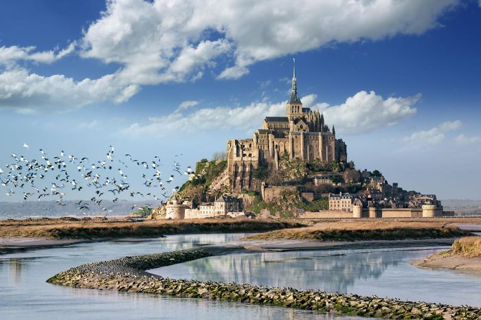 Fairy tale village with a historic castle on a rocky island surrounded by water and flocks of birds in flight.