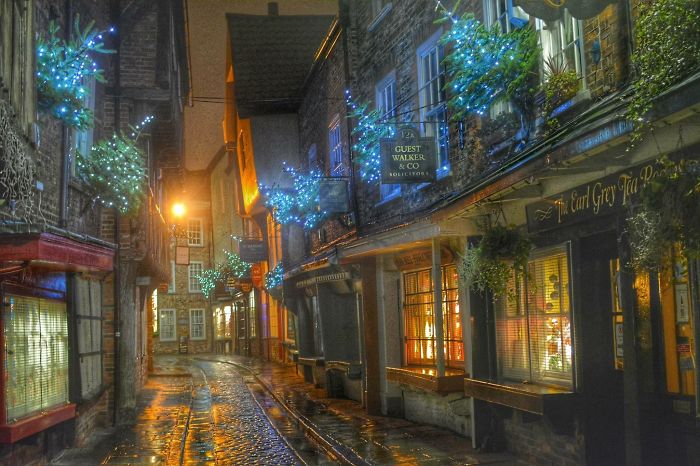 Charming fairy tale village street at night with illuminated shops and cobblestone path, adorned with festive blue lights.