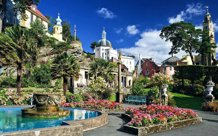 Fairy tale village with colorful buildings, lush gardens, and a fountain under a clear blue sky.