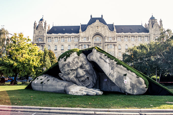 Large amazing sculpture of a giant man appearing to break through the ground covered with moss in an urban park setting.