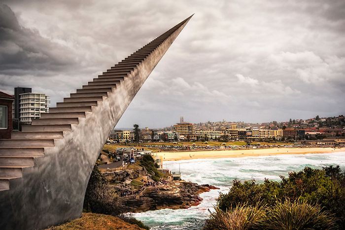 Steel staircase sculpture extending toward cloudy sky overlooking a coastal city and beach, an amazing sculpture in the world.