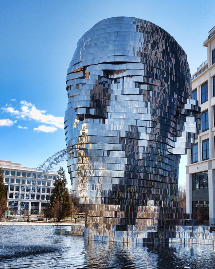 Reflective metal sculpture of a large human head with water spouting from the mouth in an urban outdoor setting.