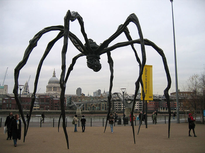 Large black spider sculpture with long legs displayed outdoors near a river in a city, one of the most amazing sculptures.