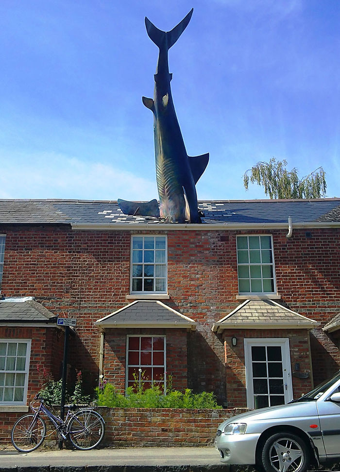 Sculpture of a shark appearing to crash through the roof of a brick house under a clear blue sky.
