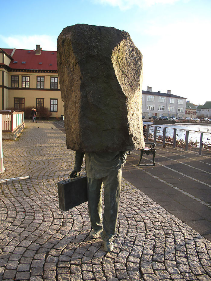 Bronze sculpture of a person with a briefcase and a large rock covering their head on a cobblestone path.
